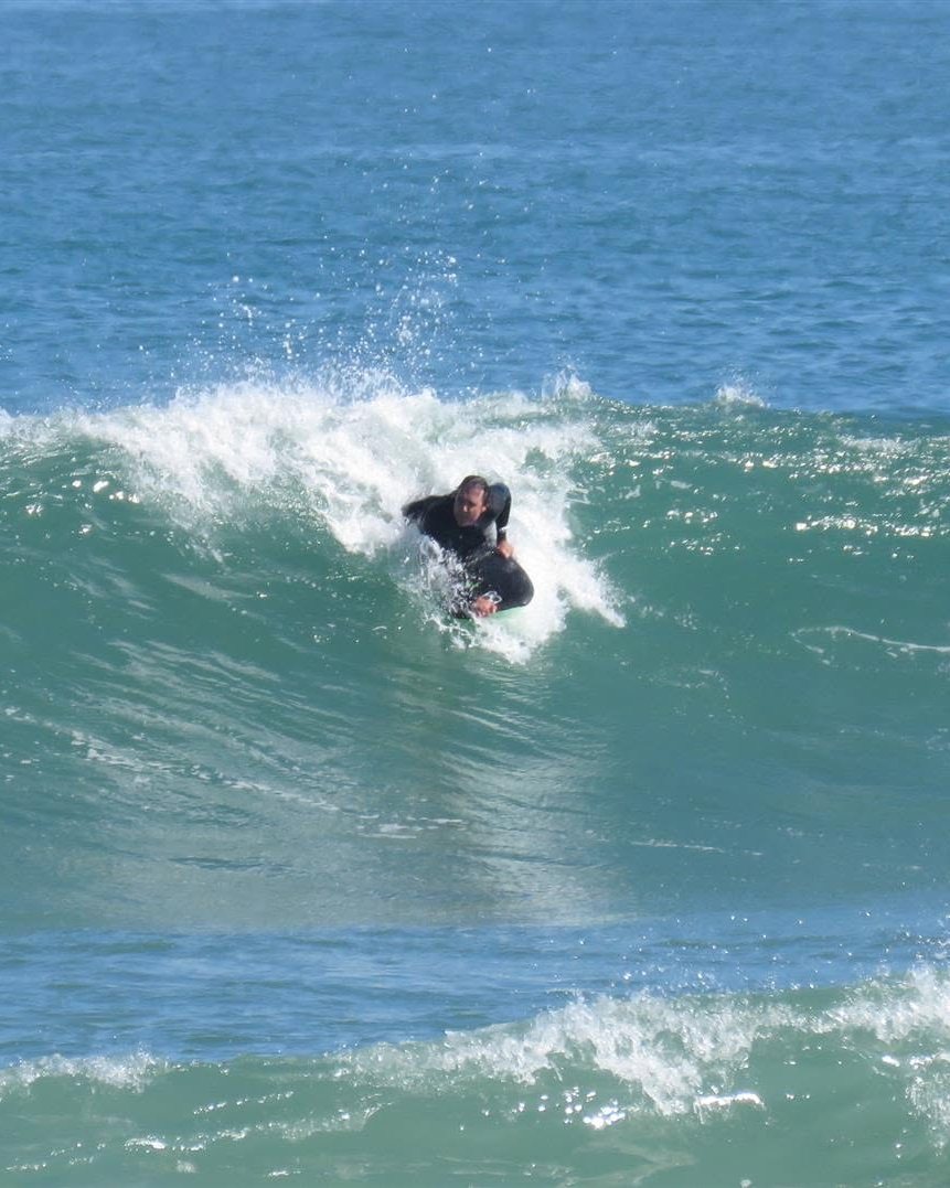 Edrich in a black wetsuit riding a bodyboard down the face of a breaking green wave